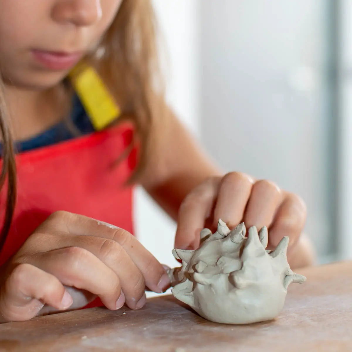 A child in a red apron molds clay on wood, building creativity at GLINARIJE NA LONTRGU: Poletna mala šola gline za otroke – AKONTACIJA by Urchic Porcelain.