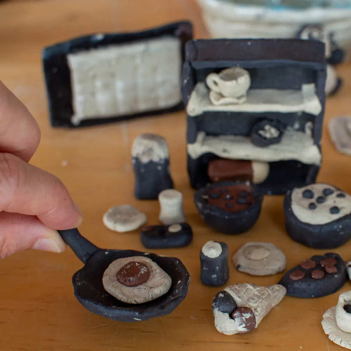 A hand holds a small frying pan with a clay egg, surrounded by mini clay kitchen items—Urchic Porcelain's GLINARIJE NA LONTRGU: Poletna mala šola gline za otroke - AKONTACIJA is perfect for kids to develop fine motor and creative skills at a summer pottery workshop.