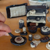 A hand holds a small frying pan with a clay egg, surrounded by mini clay kitchen items—Urchic Porcelain's GLINARIJE NA LONTRGU: Poletna mala šola gline za otroke - AKONTACIJA is perfect for kids to develop fine motor and creative skills at a summer pottery workshop.