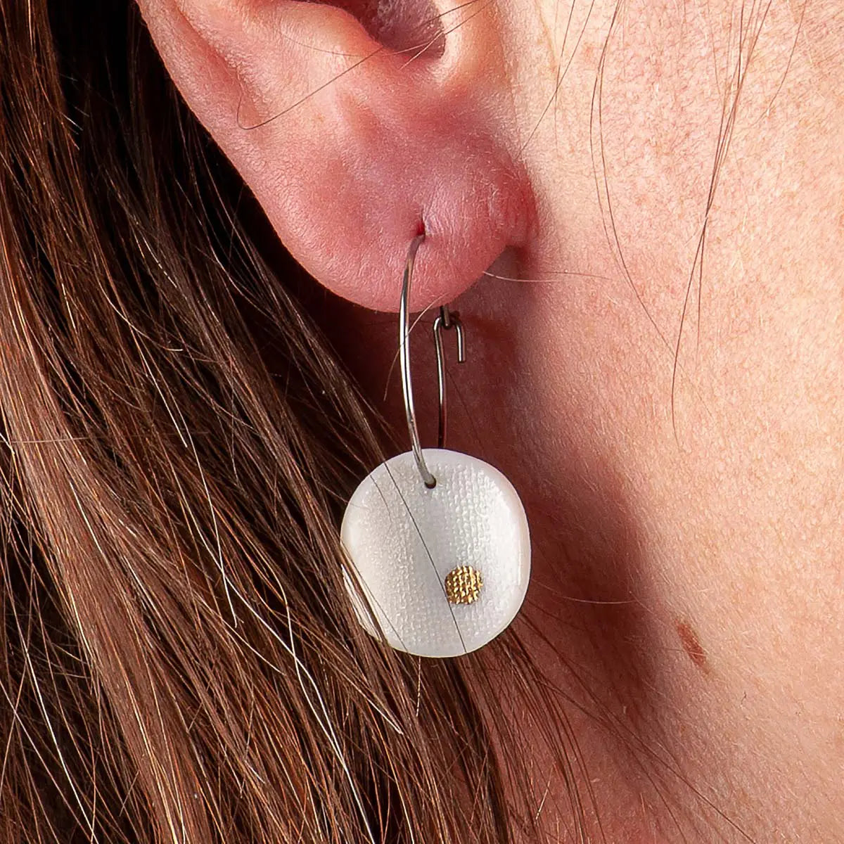 Close-up of a person's ear adorned with elegant dangling earrings, showcasing the Orion Porcelain Earrings by Urchic Porcelain, featuring a silver hoop with a white, textured disk accented by minimalist gold dots.